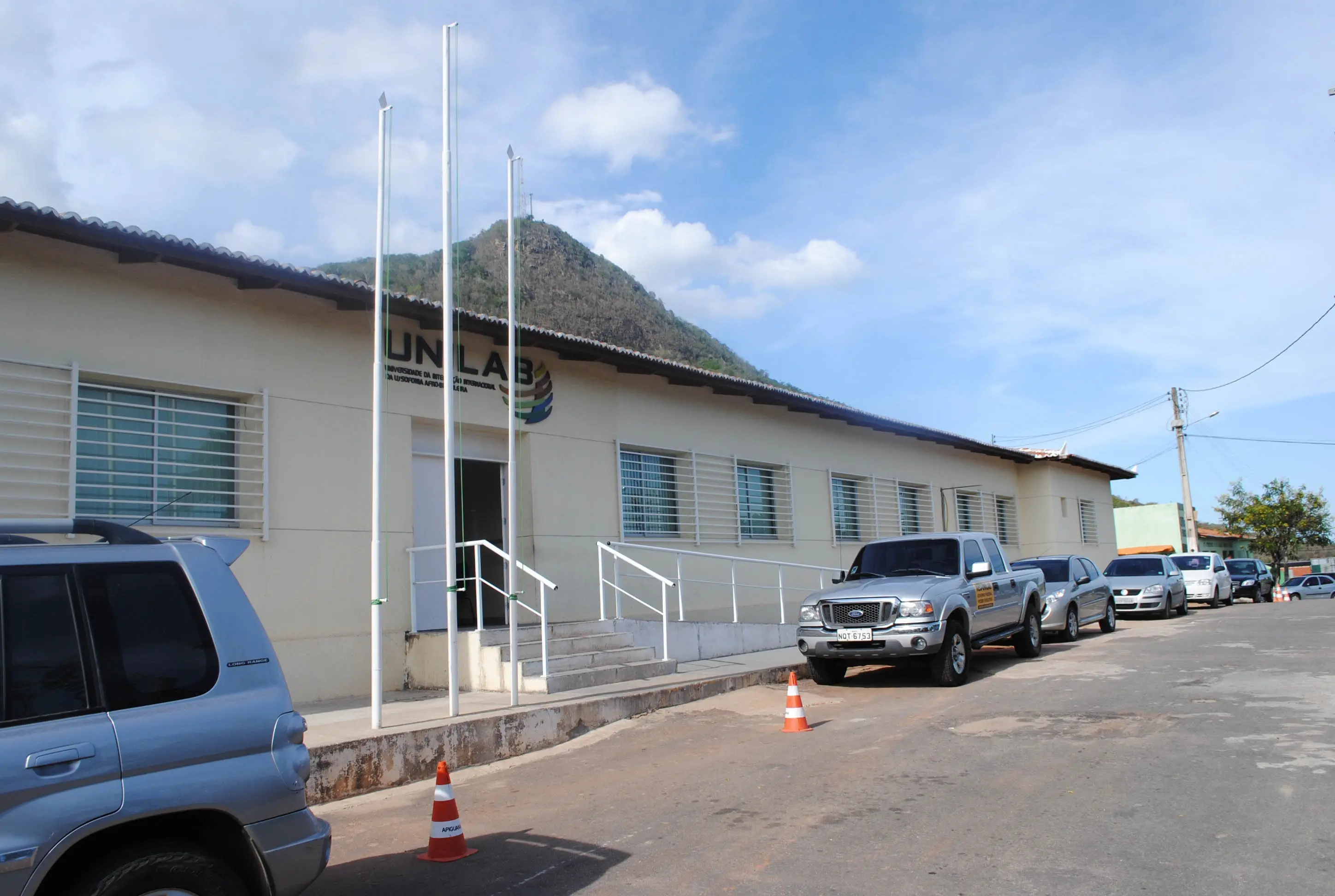 Fachada da UNILAB, prédio bege com janelas gradeadas e rampas de acesso. Carros estacionados na rua e um morro ao fundo sob céu claro com nuvens.