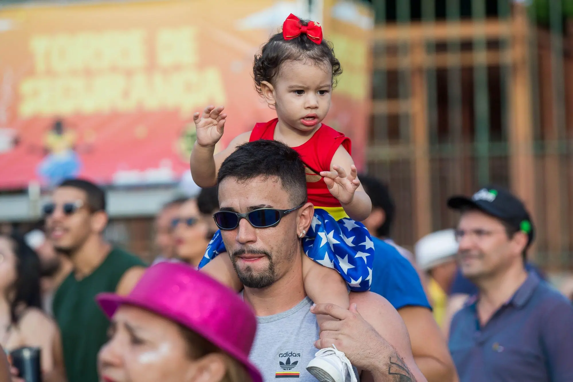Um homem com óculos escuros e barba carrega uma menina pequena fantasiada de Mulher-Maravilha nos ombros, em meio a uma multidão festiva ao ar livre.