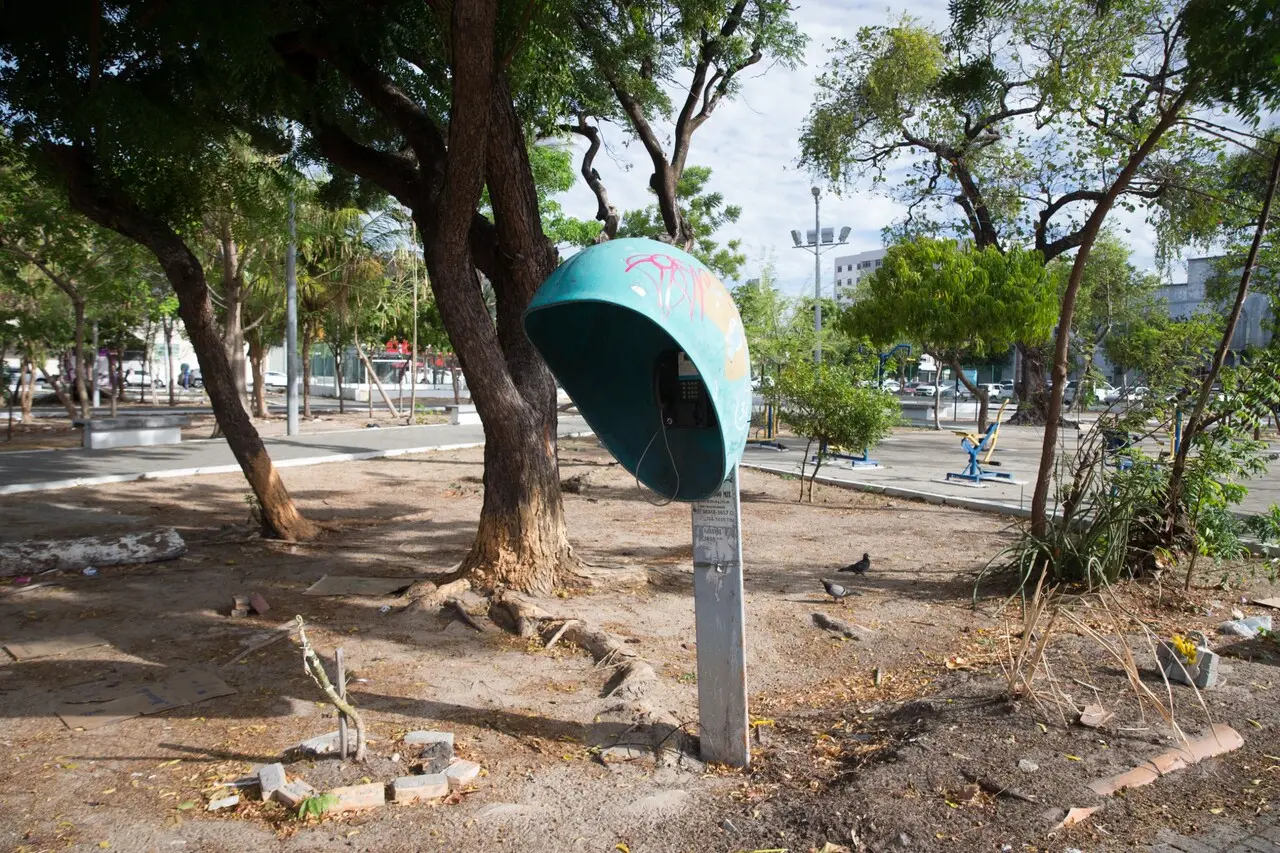 Orelhão de telefone público antigo, com cobertura azul e grafites, instalado em uma praça arborizada. Ao redor há árvores, chão de terra com folhas secas e, ao fundo, equipamentos de ginástica e prédios urbanos.