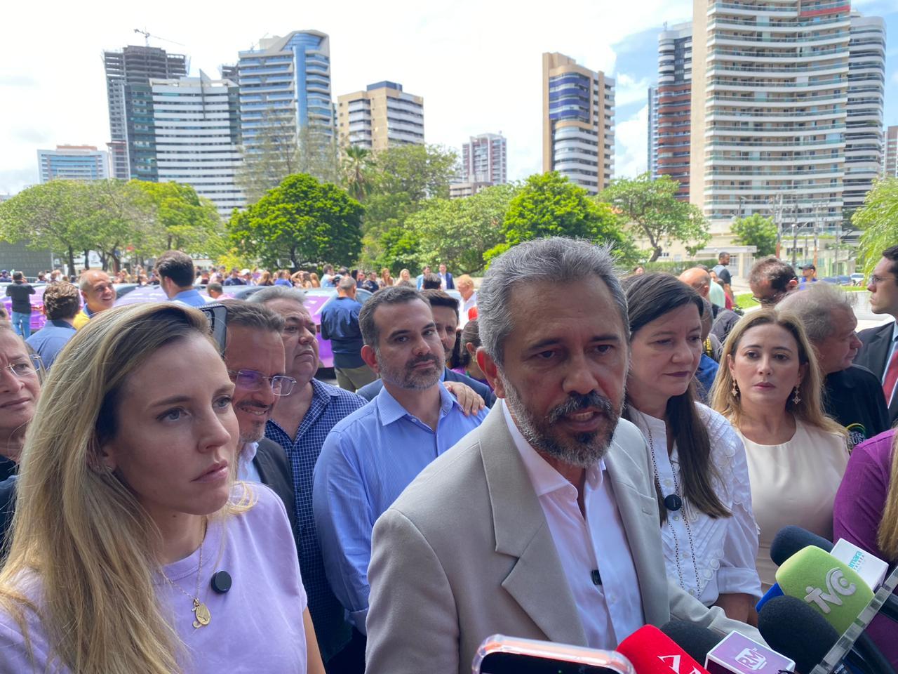 Grupo de autoridades e apoiadores reunidos ao ar livre, em área urbana com prédios ao fundo; Elmano de Freitas, governador do Ceará, fala à imprensa cercado por microfones, enquanto outras pessoas o acompanham.