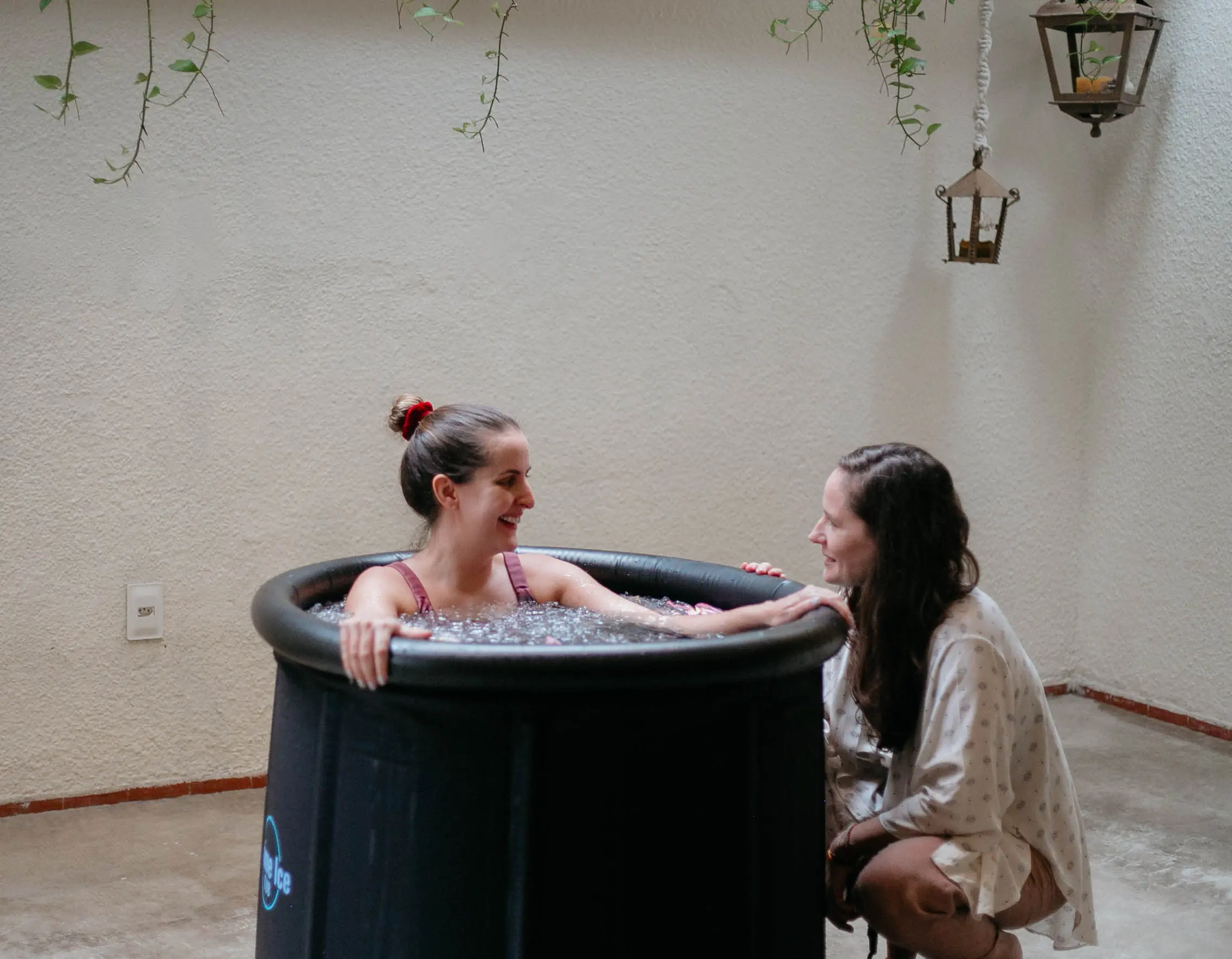 Na imagem, uma mulher sorridente submersa até os ombros em uma banheira de gelo preta e circular, localizada em um pátio externo. Ela conversa com outra mulher que está agachada ao seu lado, oferecendo apoio. O ambiente tem paredes claras, algumas plantas pendentes no topo e lanternas rústicas penduradas à direita.