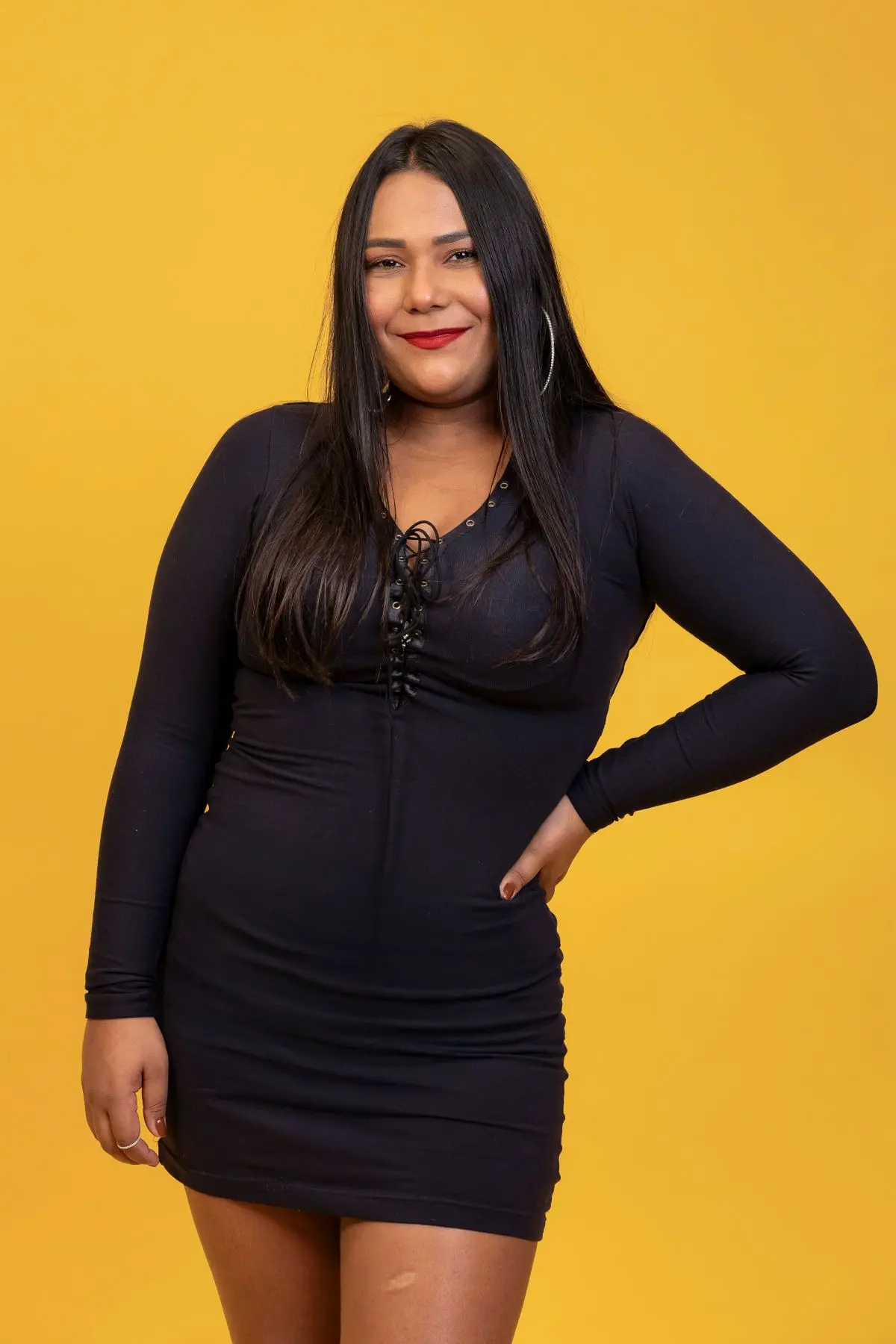 Mulher jovem e sorridente com cabelo longo e escuro, usando um vestido preto justo, em frente a um fundo amarelo vibrante.