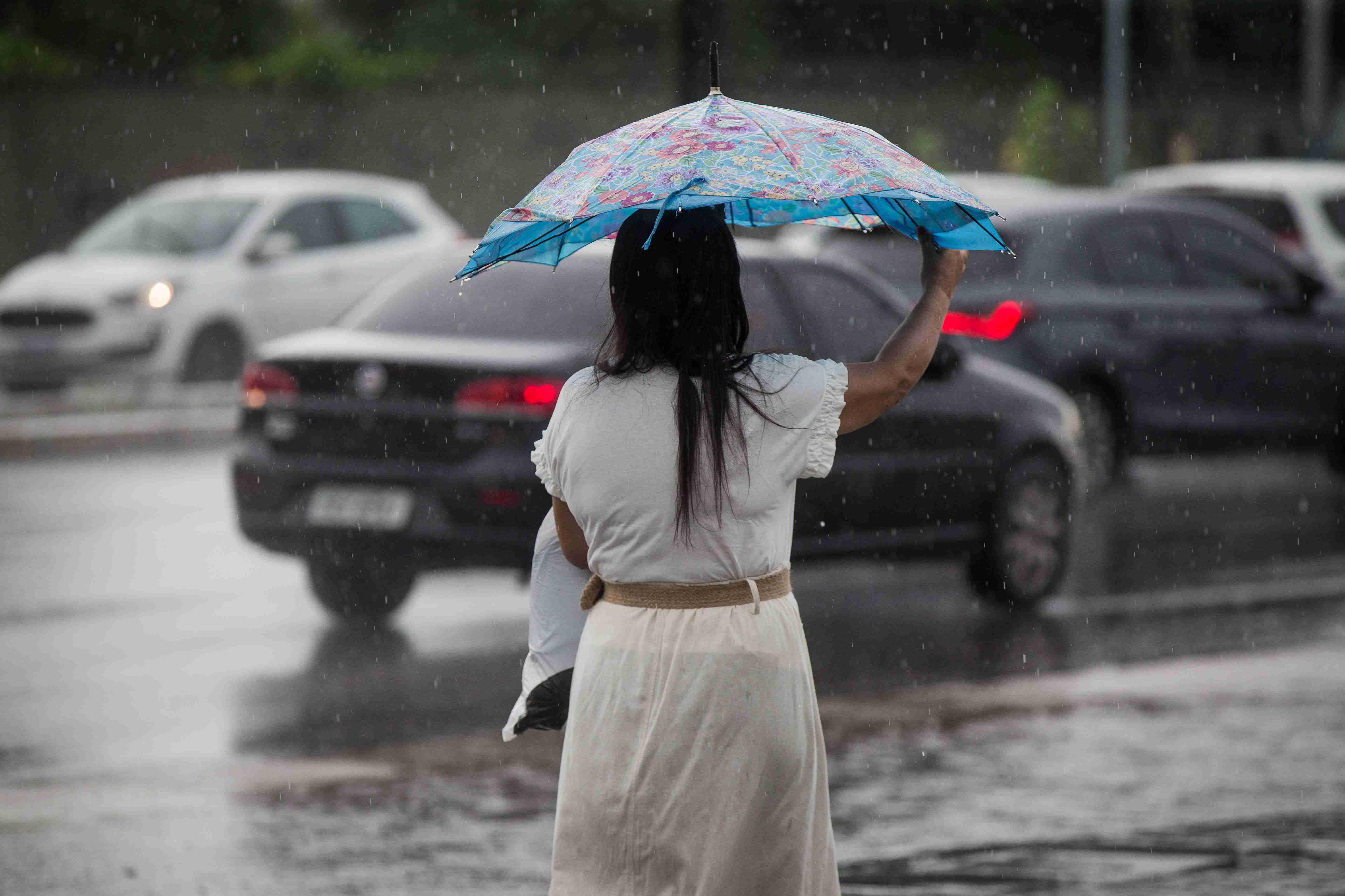 Pessoa de costas segurando um guarda-chuva azul estampado em rua molhada sob chuva leve, com carros ao fundo.