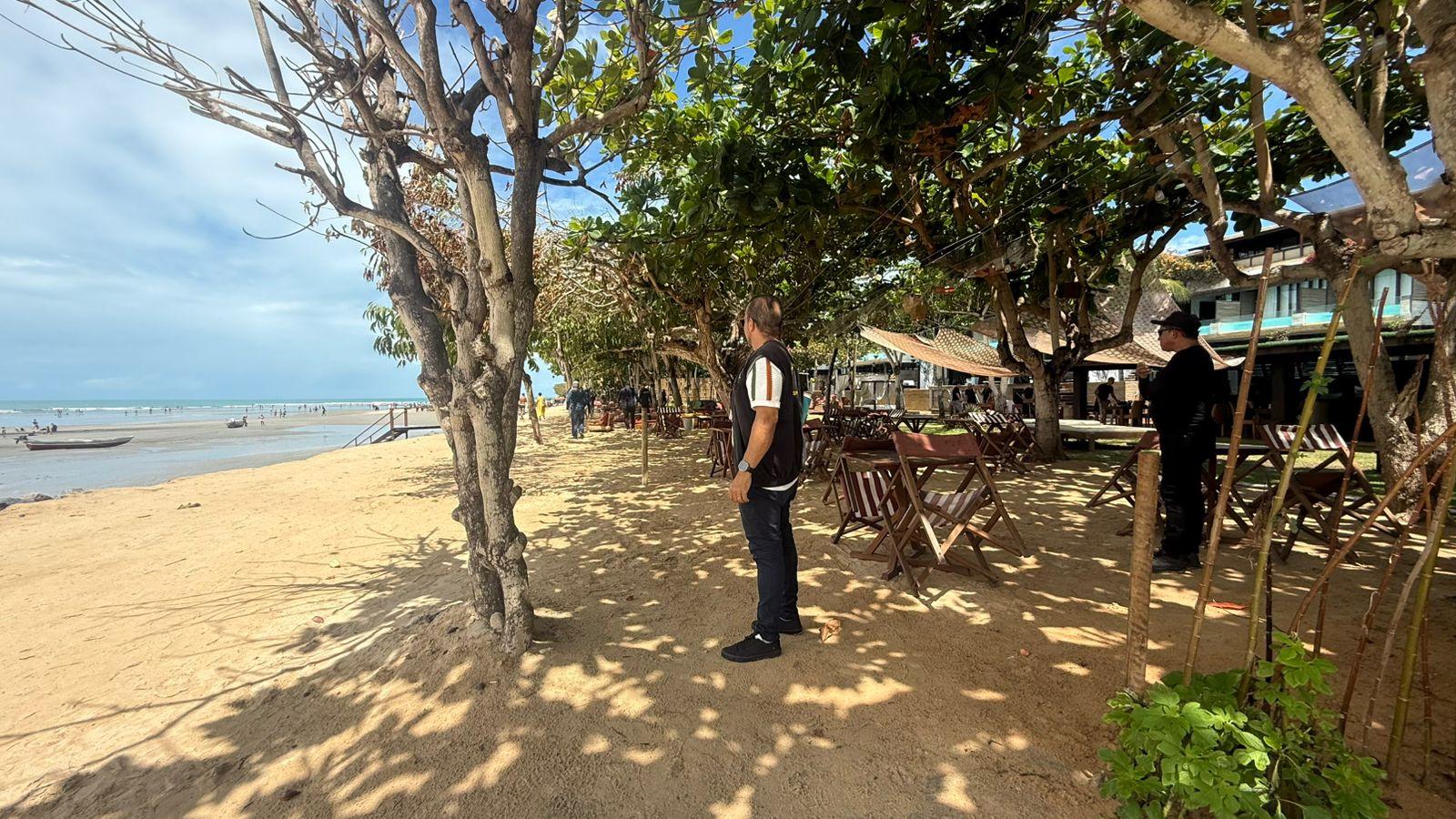 Homem em restaurante à beira-mar com mesas sob a sombra de árvores, em uma praia movimentada com pessoas e barcos no fundo.