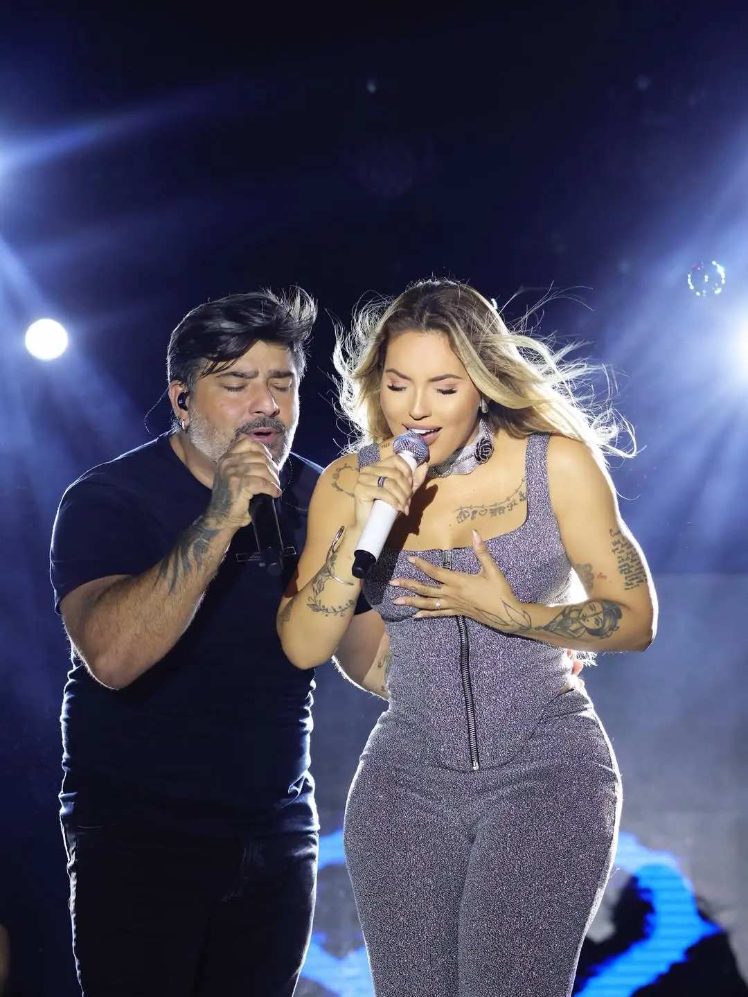 Na imagem, fotografia em plano médio de dois cantores em um show. À esquerda, um homem de pele clara e cabelos escuros, vestindo uma camiseta preta, canta com os olhos fechados e expressão emocional. À direita, a cantora Virginia Fonseca aparece com cabelos loiros soltos ao vento, usando um conjunto de top e calça brilhantes em tom lilás com zíper frontal. Ambos seguram microfones e demonstram grande sintonia. O fundo é escuro, iluminado por fortes refletores brancos que criam um efeito de aura e brilho ao redor dos artistas.