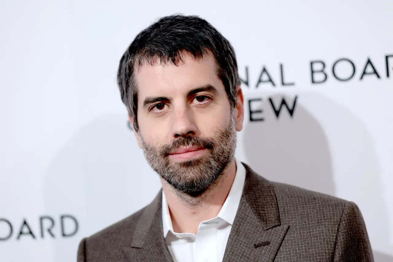 O diretor de fotografia Adolpho Veloso posa para um retrato em plano médio, vestindo um paletó cinza texturizado sobre uma camisa branca social. Ele apresenta uma expressão serena e barba por fazer, posicionado contra um fundo branco iluminado com logotipos de eventos cinematográficos.