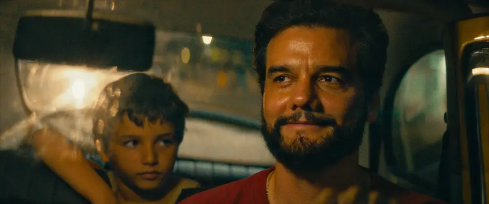 Na imagem, plano médio cinematográfico capturado de dentro de um veículo durante a noite. Em primeiro plano, à direita, está um homem (o ator Wagner Moura) com barba curta e escura, vestindo uma camiseta vermelha. Ele olha para a frente com um sorriso contido e olhos brilhantes, sugerindo um momento de quietude ou reflexão. Atrás dele, ligeiramente fora de foco no lado esquerdo da imagem, um menino observa atentamente a parte de trás da cabeça do homem ou o caminho à frente. A iluminação é quente e suave, vinda das luzes da cidade que criam um efeito de bokeh (pontos de luz desfocados) através das janelas do carro, conferindo à cena uma atmosfera nostálgica e íntima.
