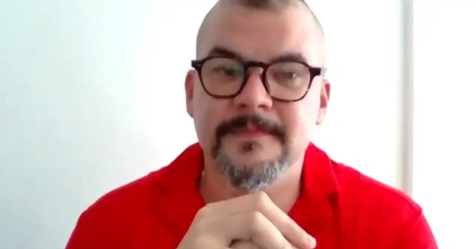 Homem branco de cabelo raspado e barba e bigode castanhos. Ele usa uma blusa de botão laranja e calça preta, e está sentado na frente de um fundo marrom.