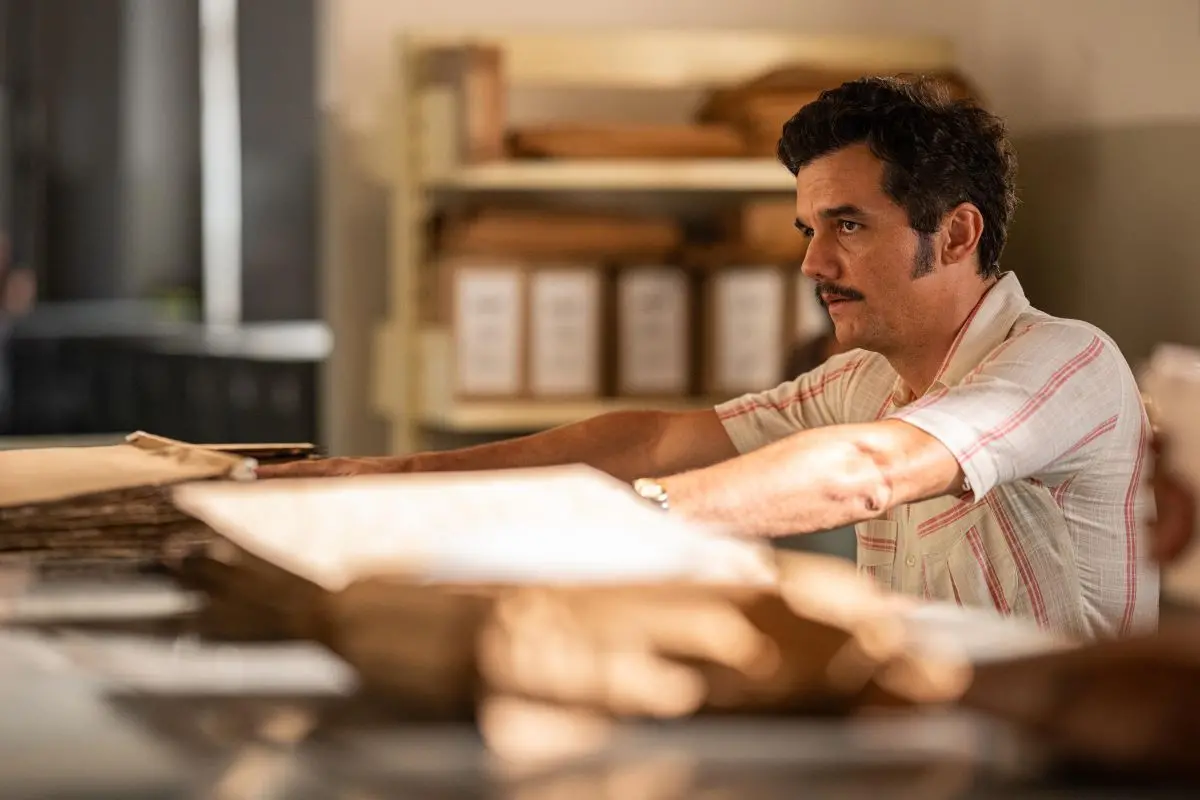 Cena de filme com homem trabalhando em ambiente de arquivo, organizando envelopes e documentos sobre uma mesa, com prateleiras ao fundo cheias de pastas catalogadas.