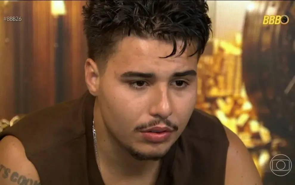 Participante do BBB 26 durante depoimento no confessionário, usando camiseta sem mangas, cenário dourado ao fundo com elementos decorativos característicos do programa.