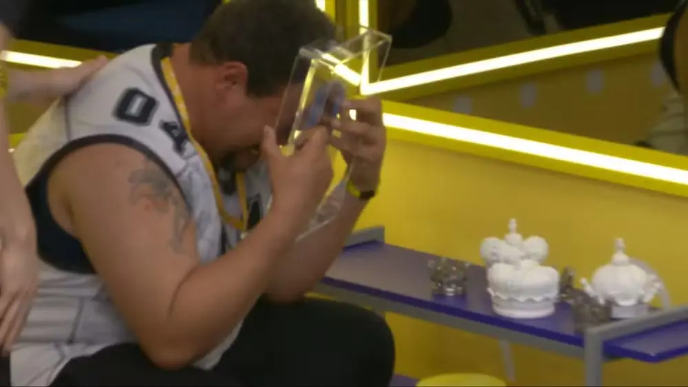 Imagem mostra Babu Santana, participante do BBB 26, chorando intensamente ao abraçar um porta-retrato com a imagem do filho logo após entrar no quarto do líder.