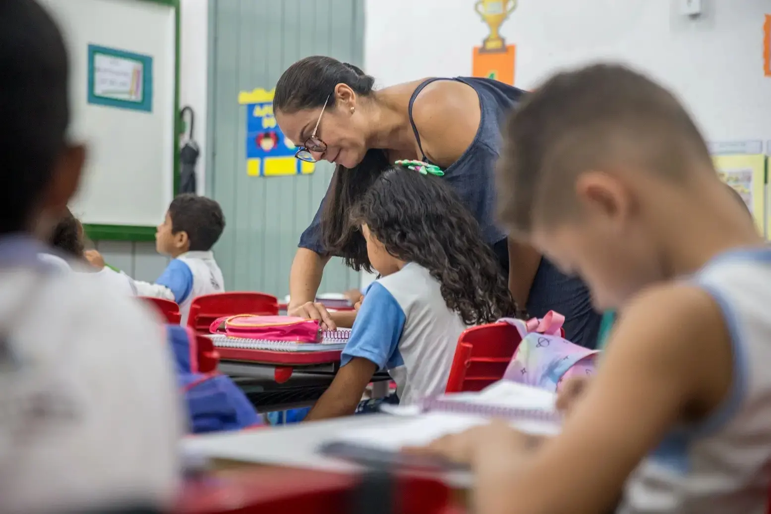 Professora da rede pública de Fortaleza auxilia aluna em sala de aula. Ambas olham para um caderno sobre a banca. Ao redor, outros estudantes uniformizados aparecem em desfoque.