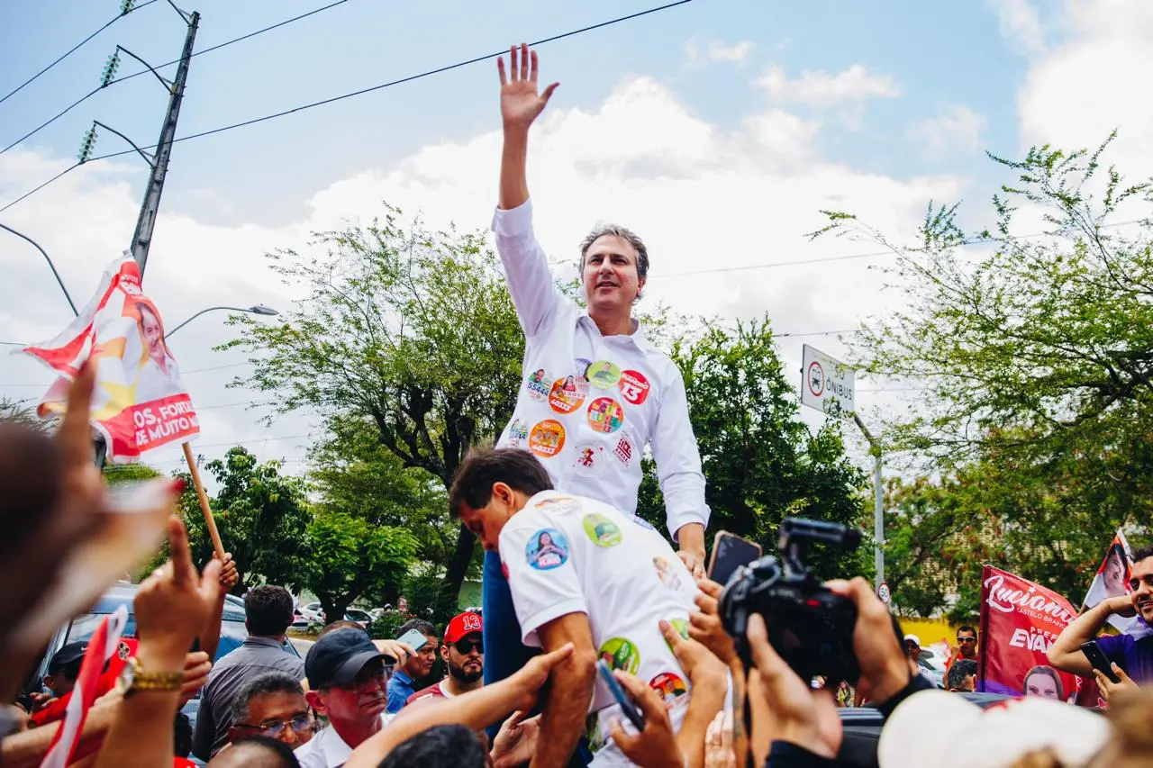 Camilo Santana, ministro da Educação, em cima de um carro ou apoiado por apoiadores, acenando com o braço levantado durante ato político em via pública; ao redor, multidão com bandeiras, celulares e câmeras registrando o momento, com árvores e céu aberto ao fundo.