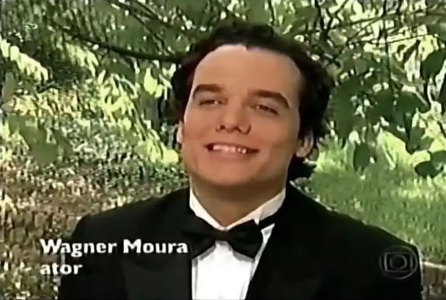 Wagner Moura, usando smoking, diante de um fundo verde. ele está sorrindo e na tela aperece o caracter WAGNER MOURA - ATOR.
