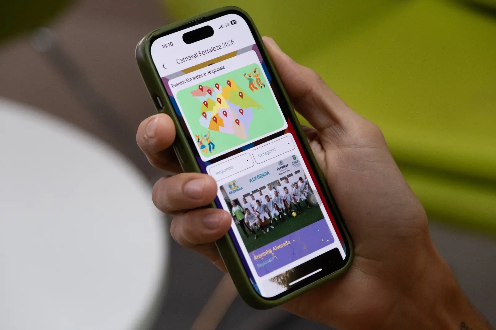 Na imagem, uma mão segura um iPhone com capa verde-oliva, exibindo na tela um aplicativo intitulado 