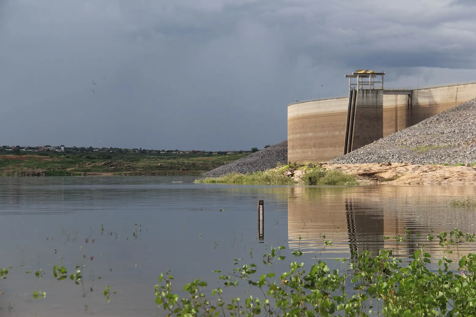 Vista ampla do Açude Castanhão com águas calmas e vegetação aquática em primeiro plano. À direita, a imponente estrutura de concreto da barragem sob um céu carregado de nuvens escuras e pesadas de chuva.