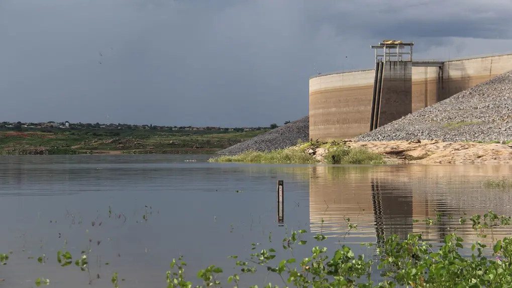 Vista ampla do Açude Castanhão com águas calmas e vegetação aquática em primeiro plano. À direita, a imponente estrutura de concreto da barragem sob um céu carregado de nuvens escuras e pesadas de chuva.