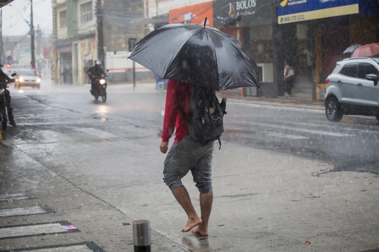 Pessoa caminha descalça pela rua sob chuva forte, segurando um guarda-chuva preto e usando mochila. Ao fundo, há carros, uma motocicleta e fachadas de comércios, com a pista molhada e a chuva caindo intensamente.