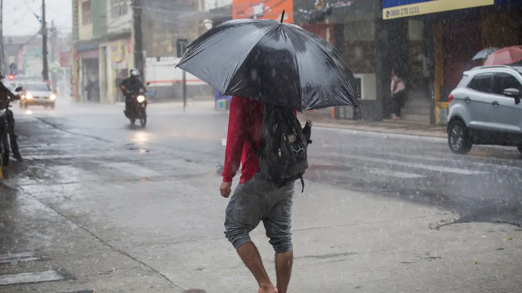 Pessoa caminha descalça pela rua sob chuva forte, segurando um guarda-chuva preto e usando mochila. Ao fundo, há carros, uma motocicleta e fachadas de comércios, com a pista molhada e a chuva caindo intensamente.