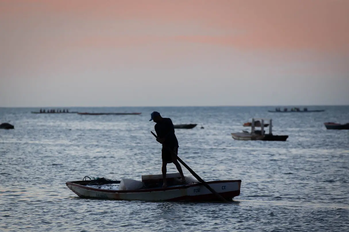 Silhueta de um pescador em pé num pequeno barco de madeira, remando no mar de Fortaleza. O céu ao fundo exibe tons suaves de rosa e laranja do entardecer, com outras embarcações distantes no horizonte.