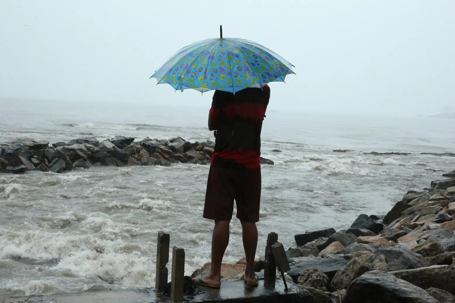 Um homem de costas segurando um guarda-chuva azul e amarelo, observa o mar agitado da praia de Iracema que bate contra rochas. O dia está nublado e chuvoso, com uma névoa densa que reduz a visibilidade ao fundo.