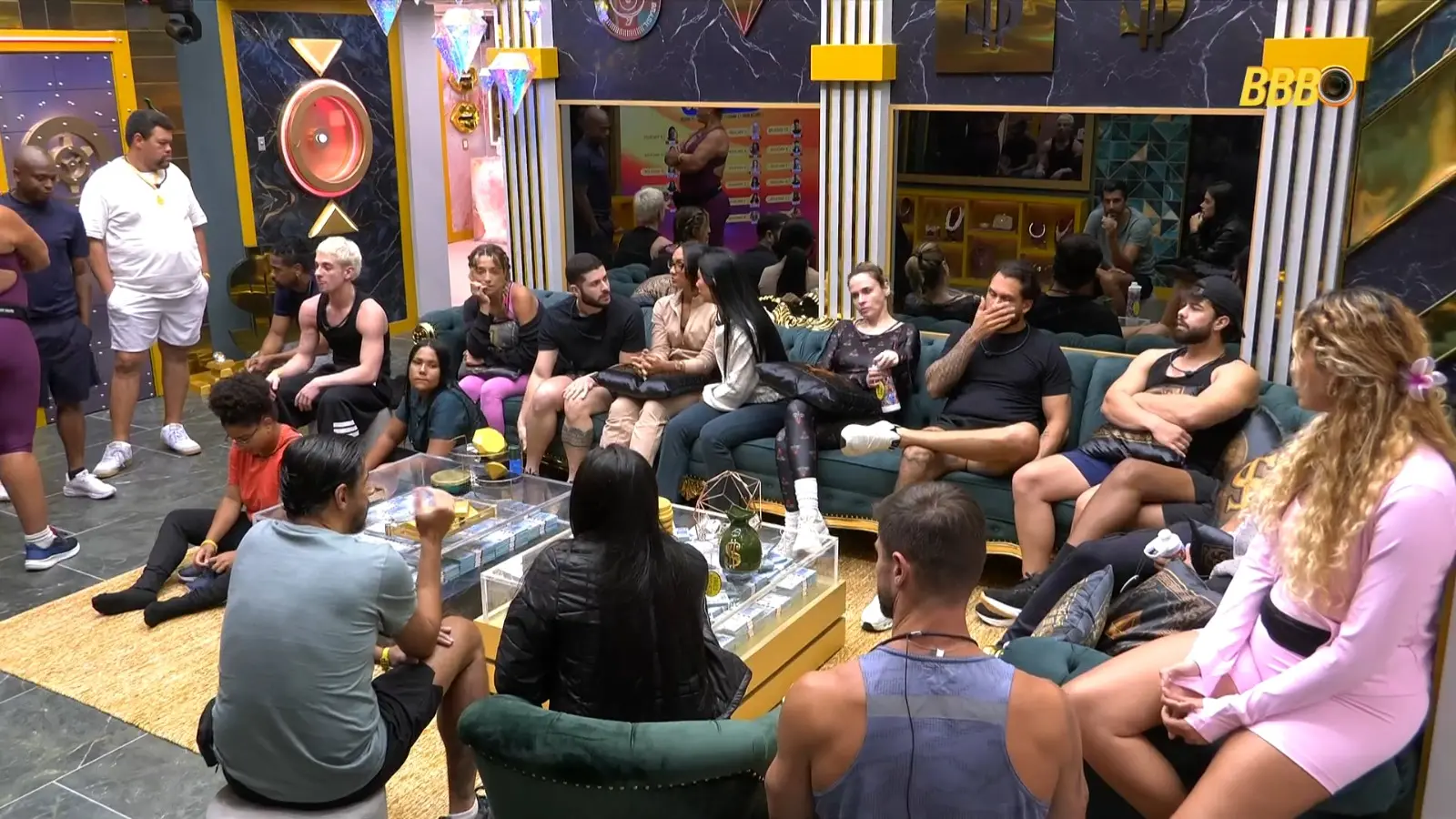 Cena interna do BBB mostrando diversos confinados sentados em um sofá e no chão, em um ambiente decorado com espelhos e itens de luxo.