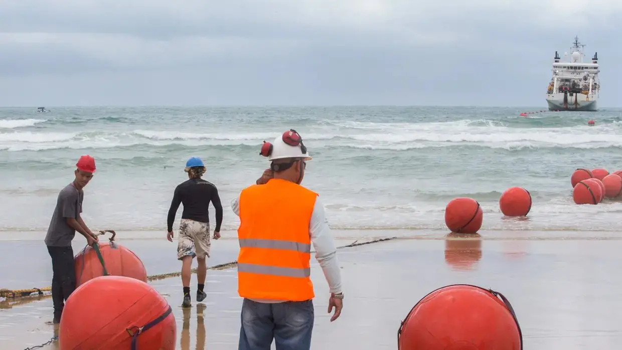 Trabalhadores usam capacetes e coletes de segurança na praia enquanto instalam boias de um cabo submarino; ao fundo, o mar e um navio de apoio responsável pela operação.
