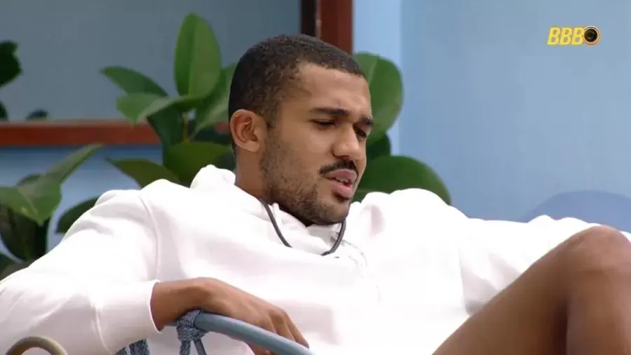 Homem sentado em área interna, usando moletom branco, com expressão pensativa enquanto olha para baixo. Ao fundo, plantas decorativas e parede azul.