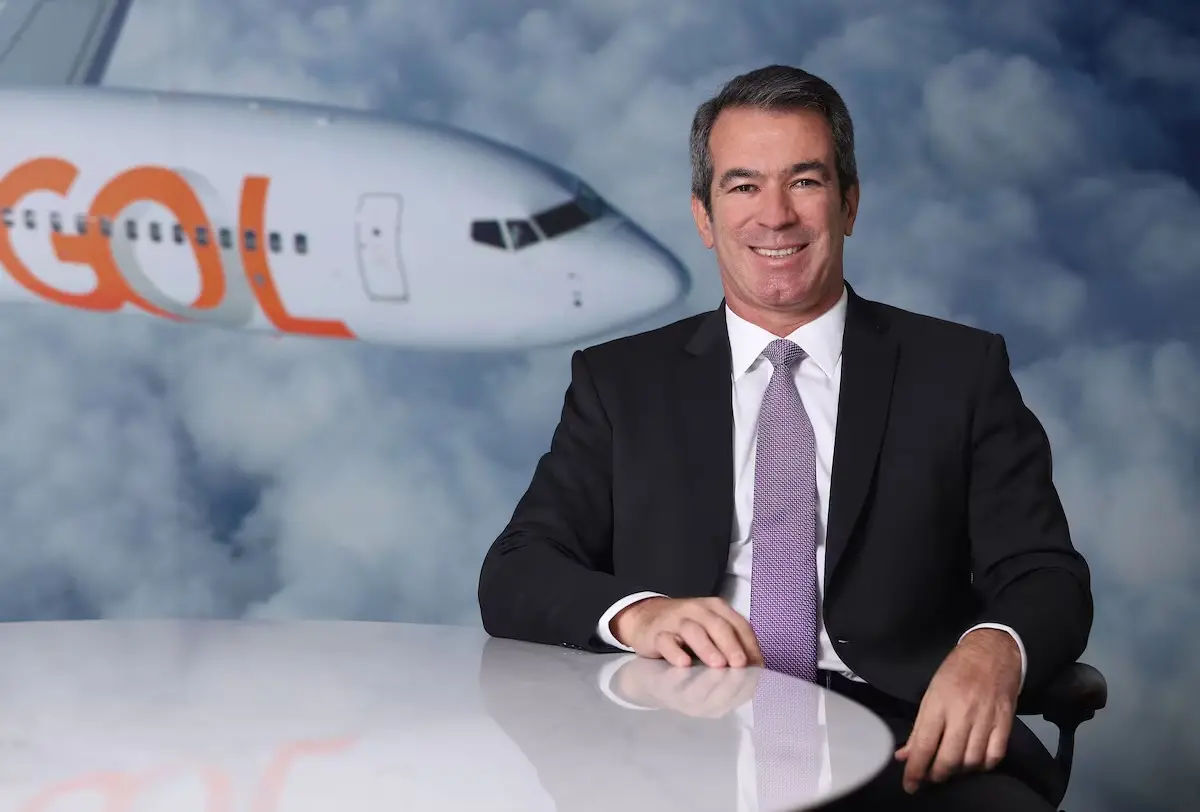 Homem de terno escuro e gravata clara sentado à mesa, sorrindo para a câmera; ao fundo, imagem de um avião com o logotipo da GOL entre nuvens, em ambiente corporativo.