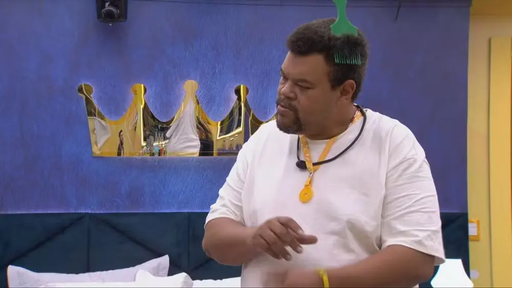 Homem em ambiente interno, vestindo camiseta branca e usando um microfone de lapela com cordão no pescoço. Ele está de perfil, fazendo um gesto com a mão, com um pente verde apoiado no cabelo. Ao fundo, há uma parede azul com um espelho em formato de coroa e uma cama com cabeceira estofada.