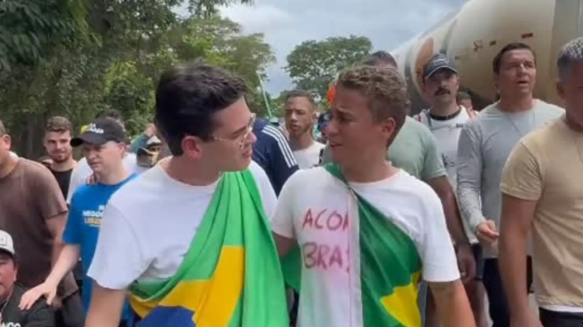 O deputado estadual Carmelo Neto e o deputado federal Nikolas Ferreira se cumprimentam em rodovia durante caminhada. Eles vestem uma camiseta branca e carregam uma bandeira do Brasil.