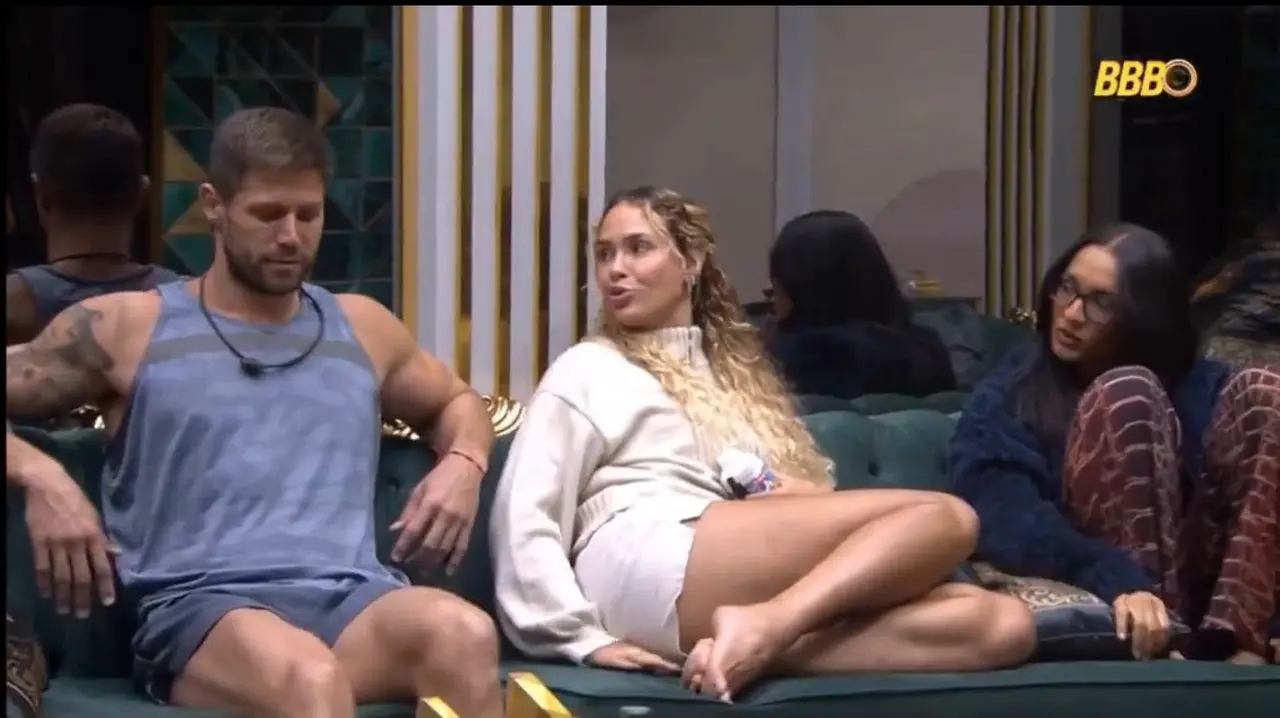 Três pessoas sentadas em um sofá. Da esquerda para direita: homem branco de cabelo baixo loiro, regata e shorts azul, com tatuagens; mulher branca de cabelo loiro, longo e ondulado, com blusa e short brancos; mulher negra de cabelos longos, lisos e pretos, com óculos, blusa longa azul e calça vermelha quadriculada.