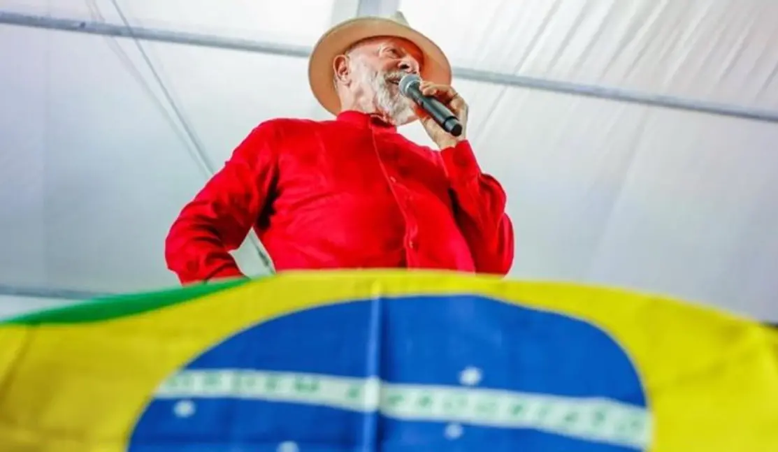 A foto mostra o presidente brasileiro Lula, vestido com uma blusa vermelha e usando um chapéu, discursando com um microfone, acima de uma bandeira do Brasil.