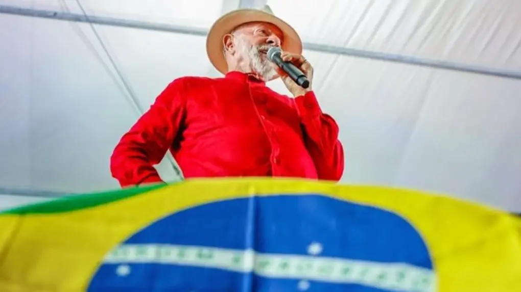 A foto mostra o presidente brasileiro Lula, vestido com uma blusa vermelha e usando um chapéu, discursando com um microfone, acima de uma bandeira do Brasil.