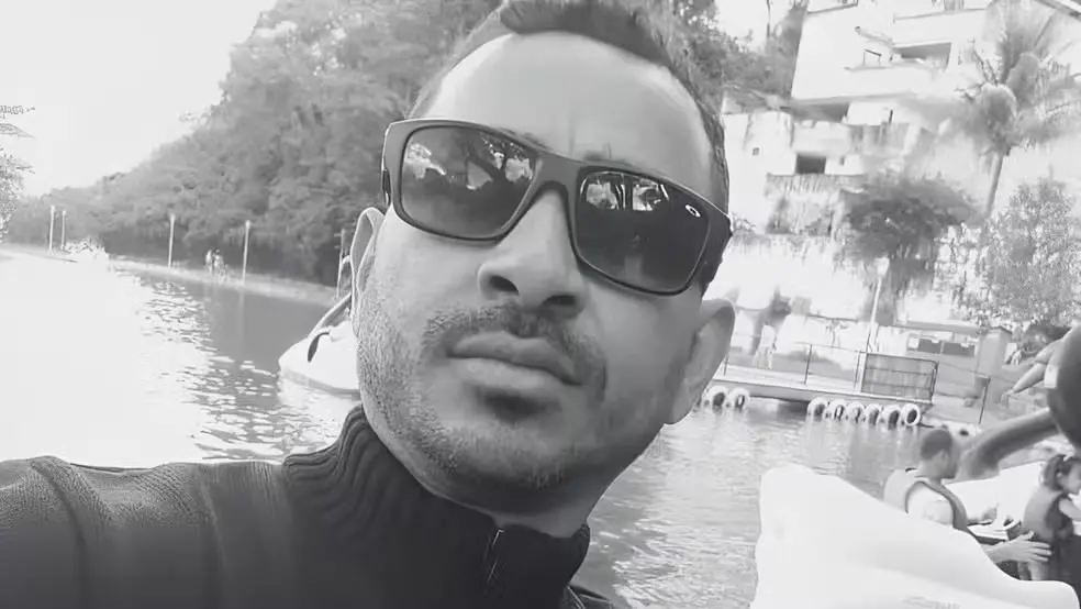 Selfie em preto e branco de um homem usando óculos escuros e jaqueta de gola alta. Ele está ao ar livre, próximo a um lago, com pessoas, pedalinhos e prédios ao fundo, além de árvores ao redor.