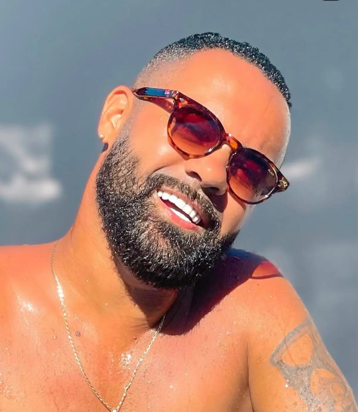 Imagem mostra o rosto de um homem negro, de barba e óculos escuros, sorrindo em um dia de sol.