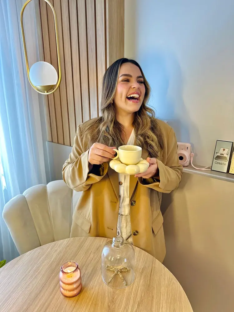 Na imagem, uma mulher jovem de pele clara e cabelos longos ondulados, com luzes loiras, sorri alegremente enquanto segura uma xícara de café. Ela veste um blazer oversized na cor bege (caramelo) sobre uma blusa clara. A xícara e o pires têm um design moderno e arredondado em tom amarelo-claro.