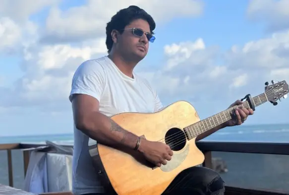 Na imagem, um homem jovem de pele clara e cabelos escuros e levemente ondulados está sentado em uma varanda ao ar livre, segurando e tocando um violão acústico de madeira clara. Ele veste uma camiseta branca básica, óculos de sol retangulares de lentes azuladas e possui uma tatuagem visível no antebraço direito. O homem olha para o lado com uma expressão serena. Ao fundo, há um mar azul sob um céu com nuvens brancas, sugerindo um ambiente litorâneo e ensolarado. A varanda possui um guarda-corpo de madeira escura.