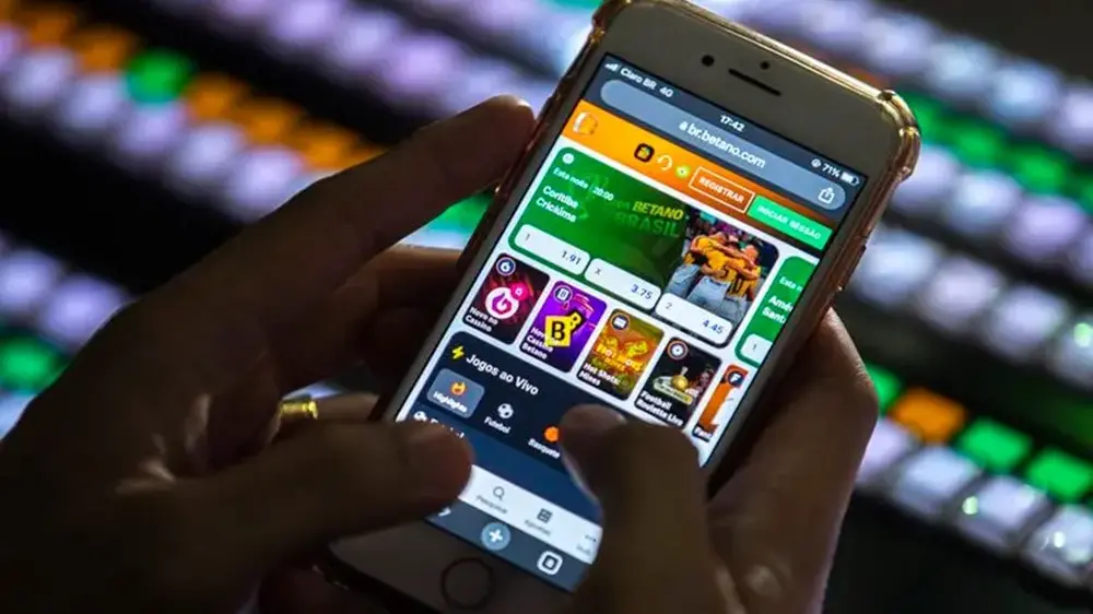 Na imagem, uma pessoa segura um smartphone com as duas mãos, exibindo na tela o site de apostas online Betano. O polegar direito está posicionado sobre a tela, indicando o uso do aplicativo ou site. A interface mostra opções de jogos ao vivo, cassino e apostas esportivas, como uma partida entre Coritiba e Criciúma. Ao fundo, fora de foco, vê-se um teclado ou painel de controle com teclas iluminadas em diversas cores (verde, laranja, azul), sugerindo um ambiente tecnológico ou de estúdio. A iluminação é baixa, destacando o brilho da tela do celular.