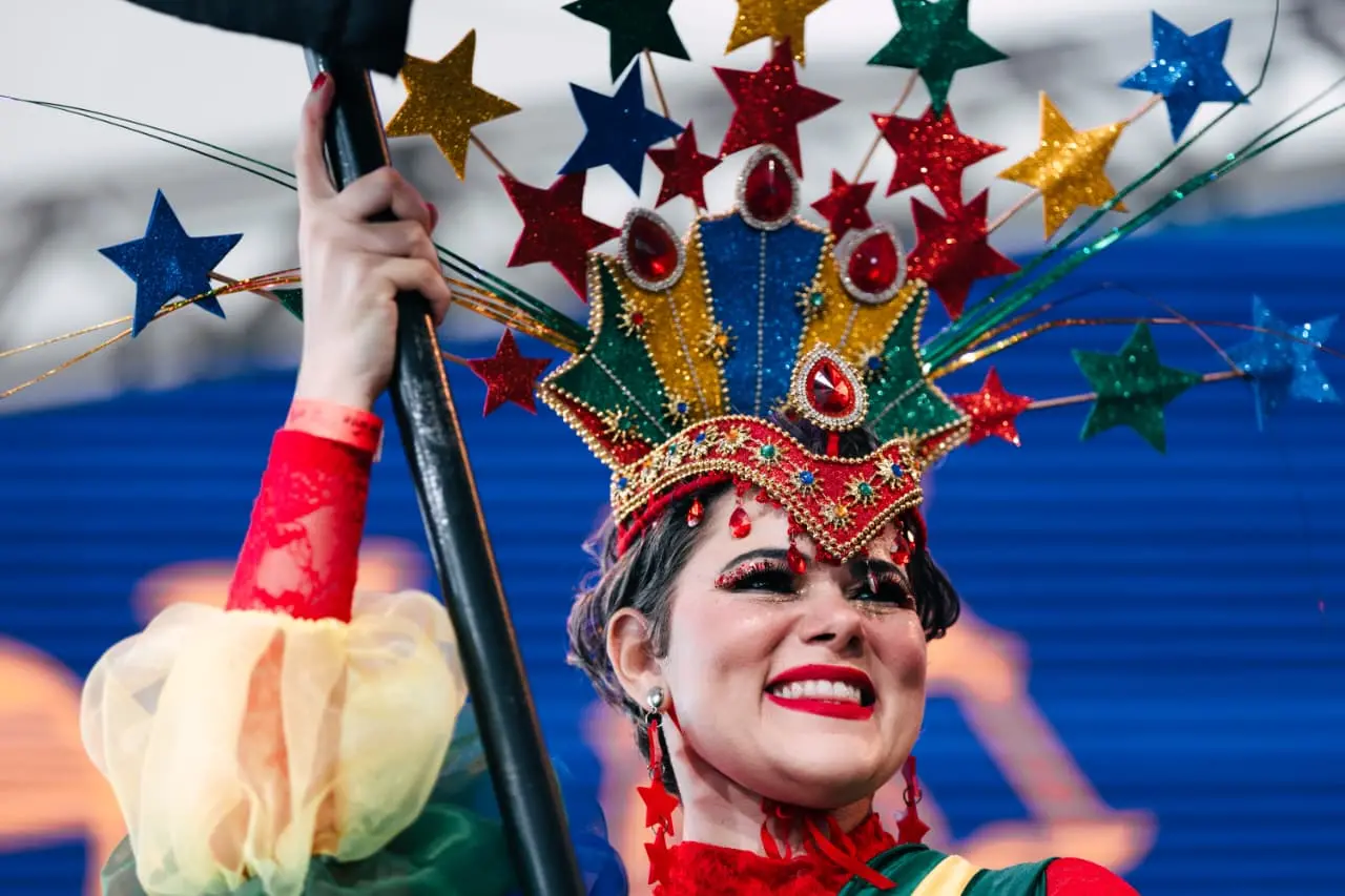Mulher maquiada e com adereço carnavalesco colorido na cabeça sorri para o público.