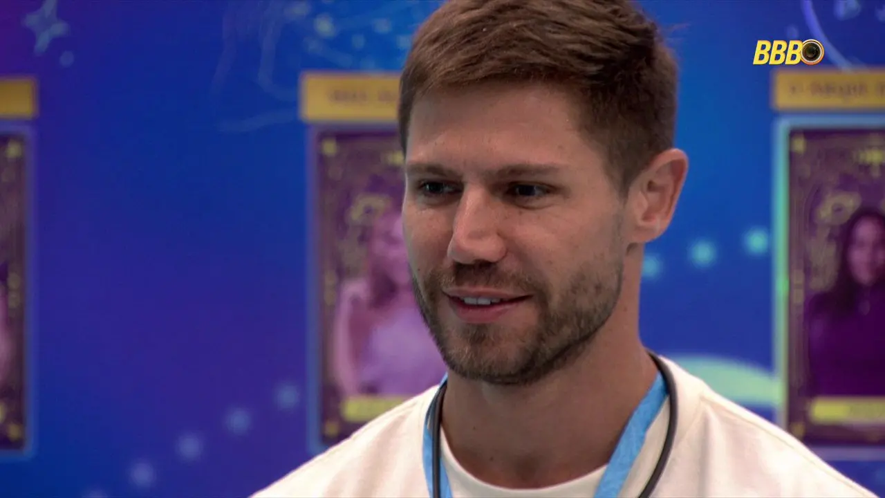 Participante em ambiente de reality show com painel azul ao fundo exibindo cards de outros competidores e logo do programa no canto superior direito.