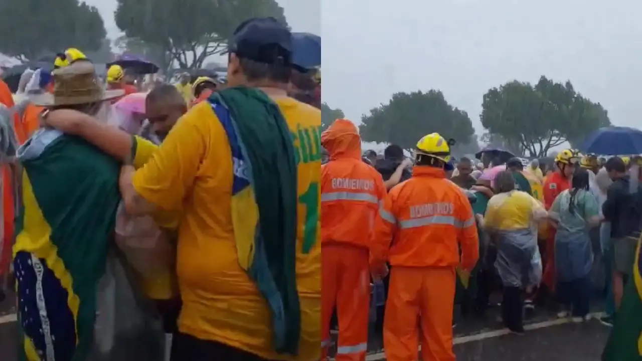 Multidão reunida sob chuva em evento ao ar livre, com pessoas usando capas de chuva e camisetas com bandeiras do Brasil, enquanto equipes de bombeiros em uniformes laranja auxiliam na organização do local.