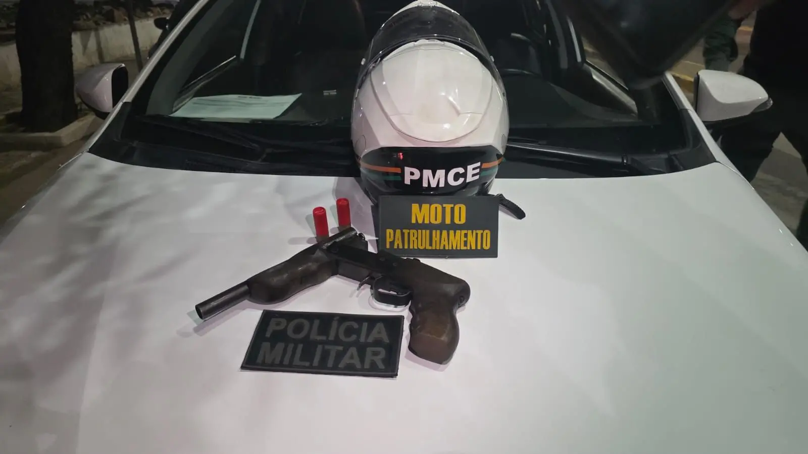 Imagem mostra arma de fogo, do tipo espingarda, em cima do capô de um carro.