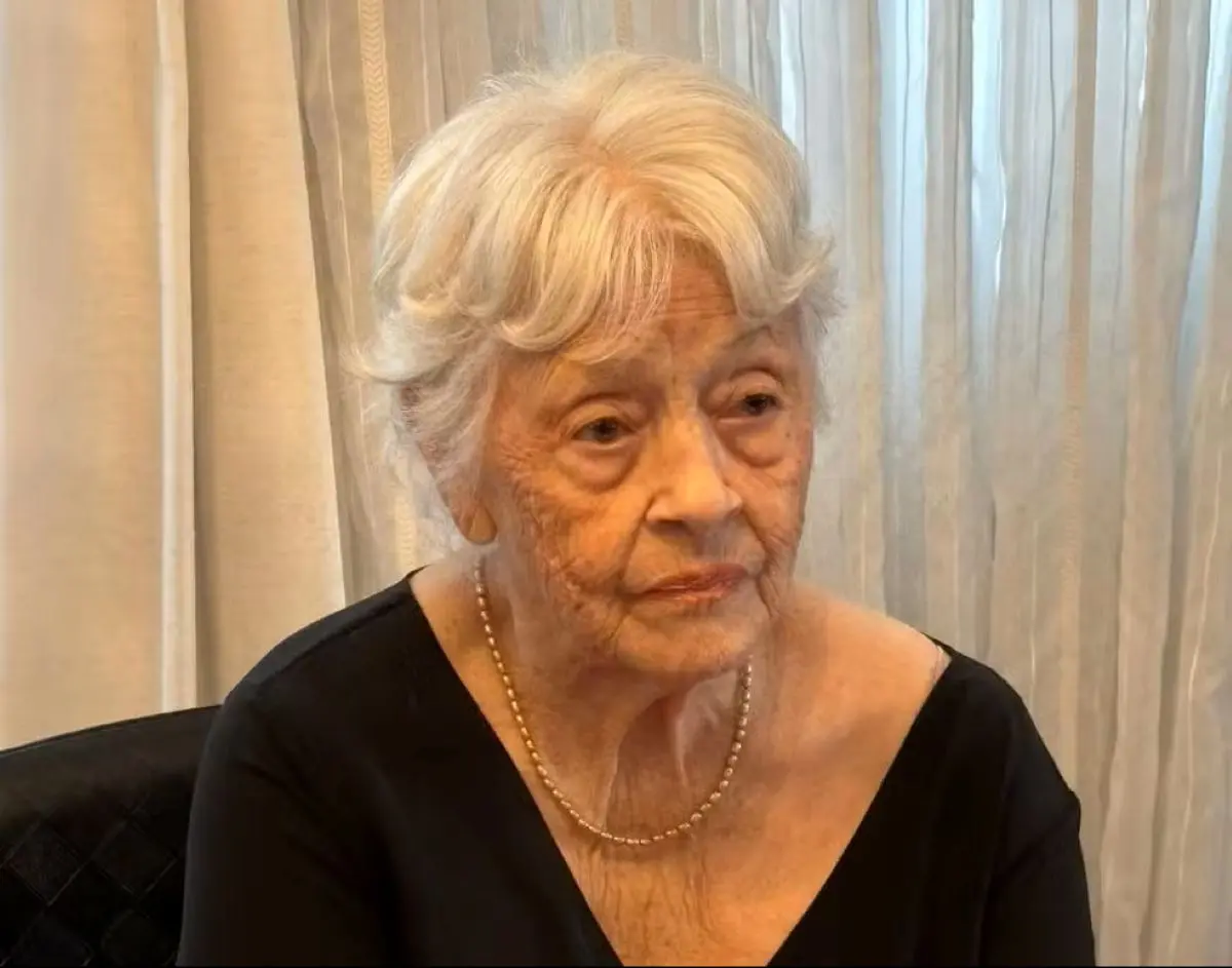 Senhora de cabelos grisalhos vestindo blusa preta e colar de pérolas, sentada em ambiente interno com cortinas claras ao fundo.