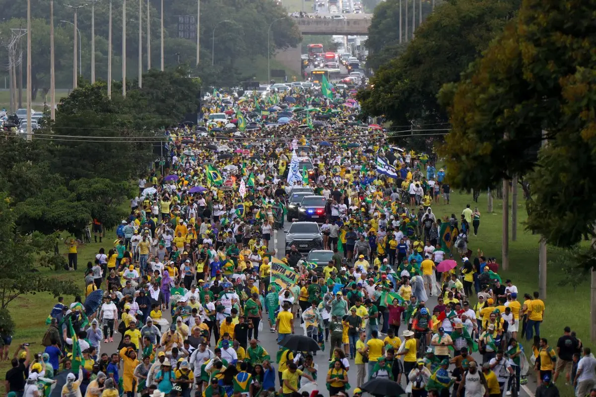 Grande multidão ocupando avenida durante manifestação com bandeiras do Brasil, roupas verde e amarelas, carros da polícia abrindo caminho e pessoas caminhando sob céu nublado em área arborizada.