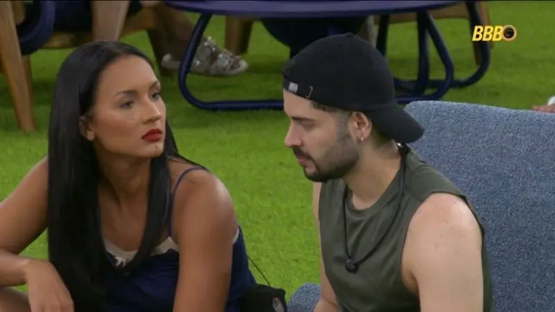 Maxiane e Marcelo, do Big Brother Brasil, sentados na grama sintética, a mulher com cabelos escuros e o homem de boné, em um momento de conversa ou reflexão.