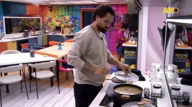 Brigido preparando uma refeição no fogão, em um ambiente de cozinha extravagante e multicolorido, do BBB 26.