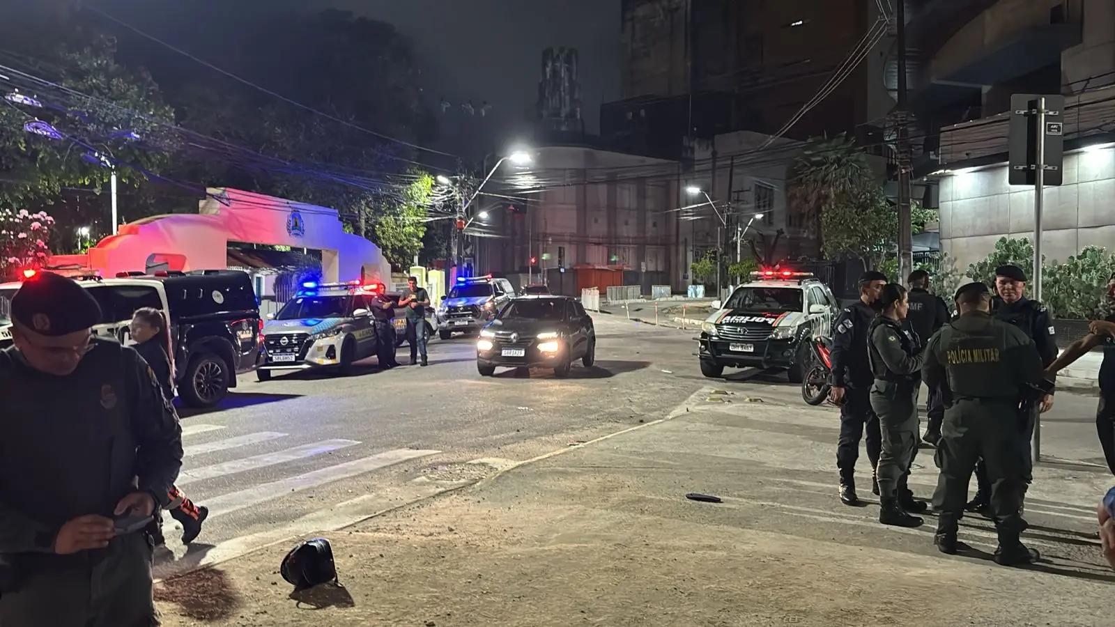 Cena noturna em uma rua urbana com várias viaturas policiais estacionadas, luzes de sirene acesas iluminando o local. Diversos agentes fardados estão reunidos e conversando próximos às viaturas, enquanto outros circulam pela área. O ambiente inclui prédios ao fundo, postes de luz e calçadas, sugerindo uma operação ou ocorrência policial em andamento.