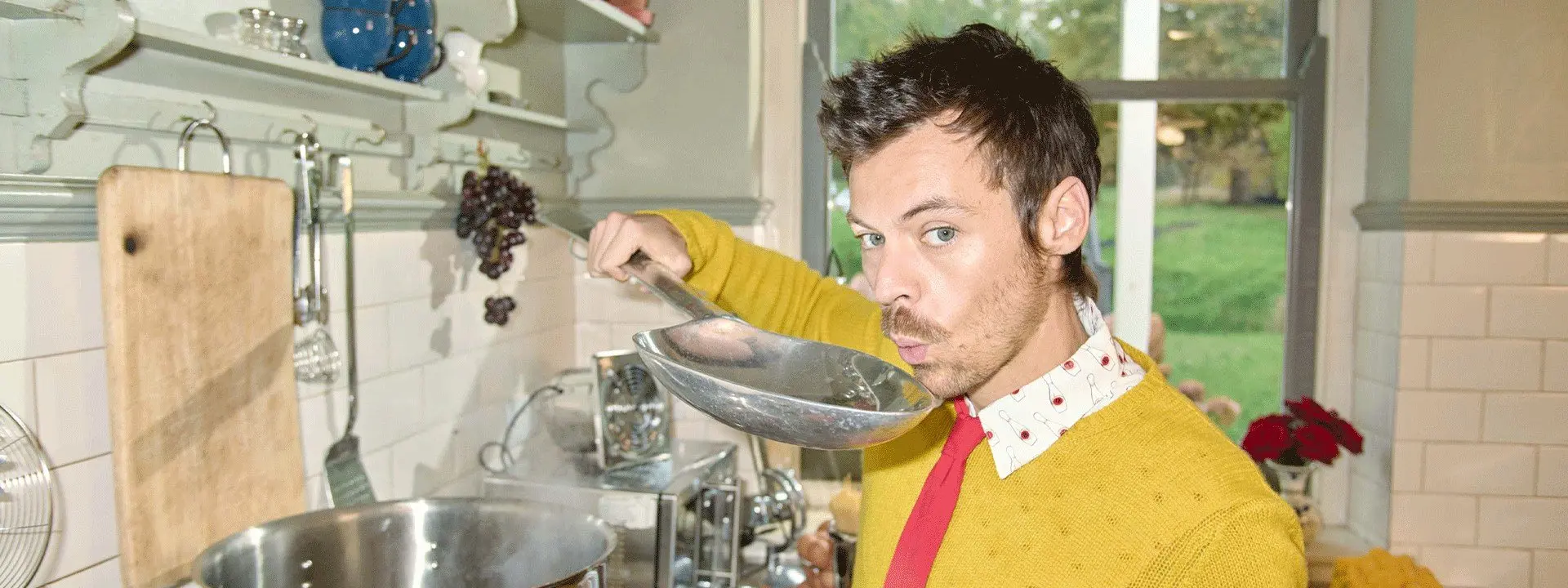 Imagem mostra cantor Harry Styles em uma cozinha, provando algo de uma concha grande com uma expressão divertida, usando um suéter amarelo e gravata vermelha.