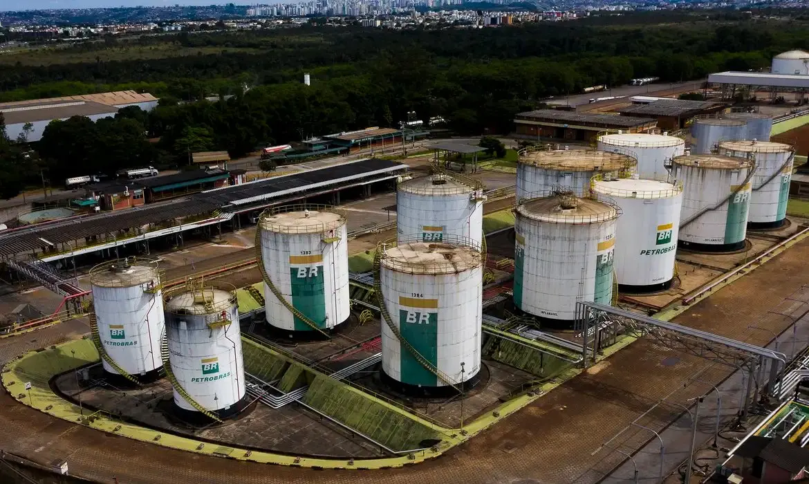 Vista aérea de múltiplos tanques de armazenamento de combustível da BR Petrobras em um terminal industrial, com área verde e cidade ao fundo.